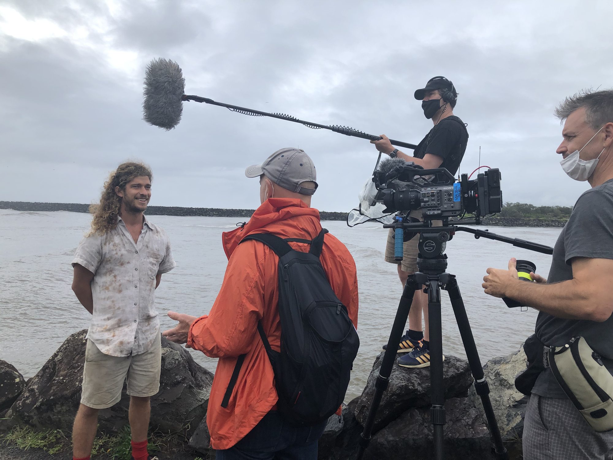 Full production crew — Sony camera on slider, boom operator, producer, and contributor on coastal rocks